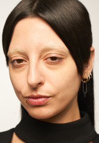 Gros plan sur une personne avec des cheveux noirs raides, des sourcils clairs, des lèvres légèrement brillantes, portant un col roulé noir et des boucles d'oreilles en or.