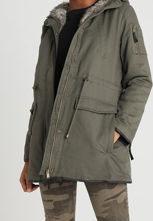 Hombre con parka verde oliva con capucha forrada de piel, cremallera frontal, bolsillos con solapa y pantalones con estampado de camuflaje.