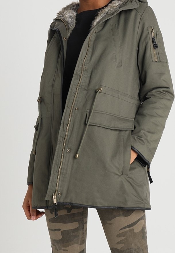Homme portant une parka vert olive avec capuche bordée de fourrure, fermeture éclair à l'avant, poches à rabat, et pantalon à motif camouflage.