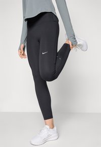 Leggings sportivi neri con una texture liscia, dotati di un logo Nike bianco sulla coscia sinistra, abbinati a una maglietta grigia a maniche lunghe e scarpe bianche.