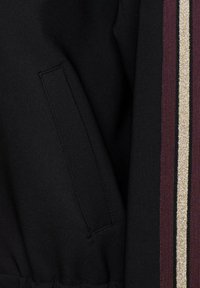 Zwarte jas van stof met een zijzak, voorzien van verticale strepen in bordeauxrood, goud en zwart. Zachte textuur met elastische taille.