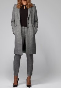 Szary w kratę garnitur z długim płaszczem i zwężanymi spodniami. Płaszcz ma podwójne zapięcie i kieszenie boczne. Pod spodem czarna bluzka, jednolity kolor.