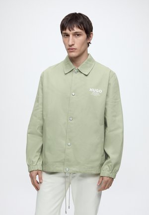 Jeune homme portant une veste boutonnée vert clair avec des poignets élastiques et le logo "HUGO" sur la poitrine, associée à un pantalon blanc, debout.