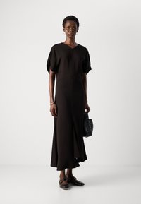 Vrouw in een zwarte midi-jurk met korte mouwen, zwarte gespschoenen, oorbellen met hoepels, een armband en een kleine zwarte gevlochten handtas.