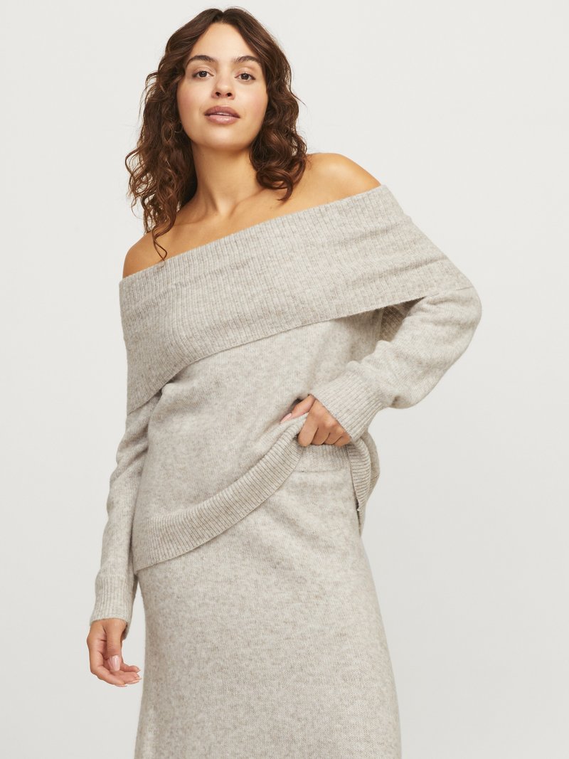 Femme portant un pull en maille côtelée beige à épaules dénudées et une jupe assortie, posant avec une main dans la poche devant un fond clair.