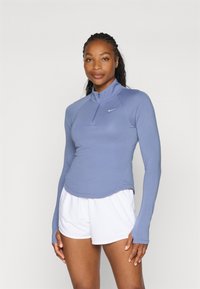Langarm-Sportoberteil in Hellblau mit einem Reißverschlusskragen, aus glattem Stoff gefertigt, kombiniert mit weißen Shorts. Nike-Logo auf der Brust.