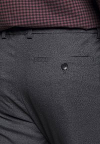 Pantaloni grigio scuro in tessuto strutturato, con una tasca posteriore dotata di bottone nero e passanti per cintura. Camicia a quadri bordeaux e neri visibile sopra.