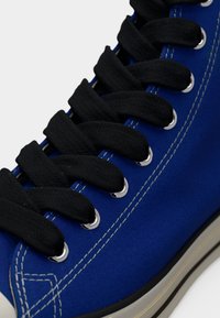 Κοντινό πλάνο μπλε καμβαδένιου αθλητικού παπουτσιού με μαύρα κορδόνια περασμένα από ασημένιες τρύπες και λευκή ραφή κατά μήκος των άκρων.