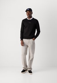 Felpa nera con colletto rotondo e un logo sottile, abbinata a jeans grigi chiari. Cappellino nero con un accent blu e sneakers nere.