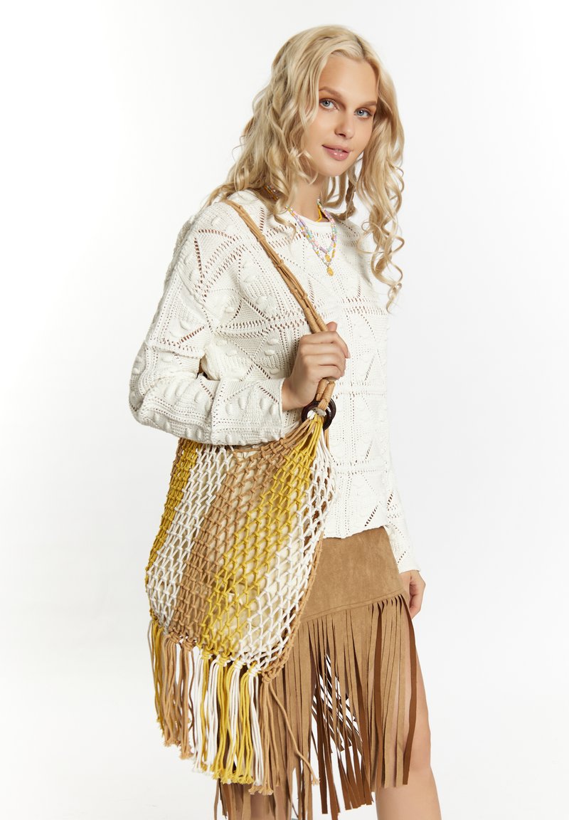 Borsa intrecciata marrone e bianca con dettagli gialli, caratterizzata da un design a rete. La modella indossa un maglione di lana bianco e una gonna in suede con frange.