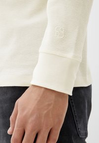 Camicia a maniche lunghe crema realizzata in tessuto a maglia waffle testurizzata. I polsini sono di un crema uniforme con un logo impresso sulla manica. Mano parzialmente visibile.
