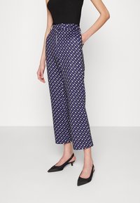 Pantalones azules con un patrón de formas geométricas en blanco y verde, de cintura alta, con bolsillos laterales, hechos de una tela suave, combinados con zapatos negros de tacón con tira en el talón.