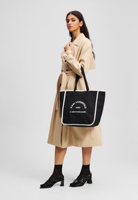 Sac fourre-tout en toile noire avec des accents blancs, portant le logo et le texte "Karl Lagerfeld Paris". Le modèle porte un trench beige et des talons noirs.