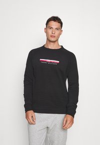 Sudadera negra de manga larga con un diseño de franjas en blanco y rojo en el pecho. Combinada con pantalones de chándal grises. Textura suave.