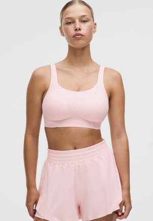 Donna che indossa un reggiseno sportivo rosa chiaro e pantaloncini sportivi vita alta abbinati, in piedi su sfondo uniforme.