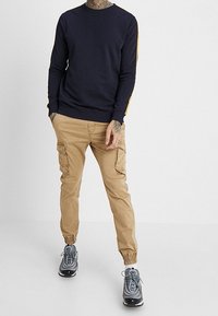 Marineblauer Langarm-Sweatshirt mit gelben Seitenstreifen, kombiniert mit beigen Cargohosen mit Seitentaschen und elastischen Bündchen. Dazu graue Sneaker getragen.