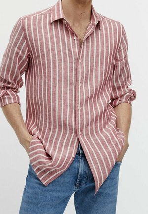 Man draagt een rood-wit verticaal gestreept overhemd met opgerolde mouwen en een blauwe spijkerbroek, handen in de zakken.