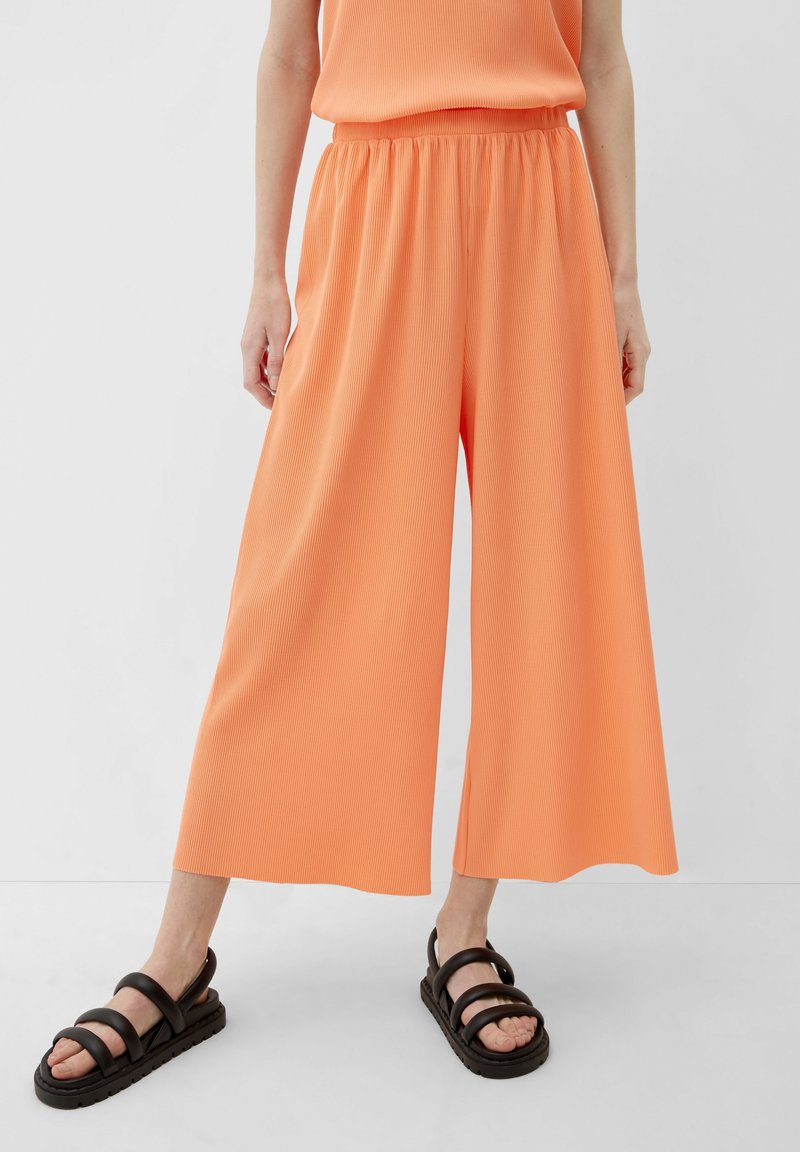 Oranje, wijde broek van ribbelstof met een elastische tailleband. G gedragen met zwarte, strap sandaal op een neutrale achtergrond.