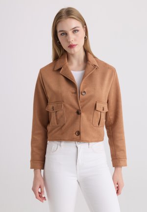 Jonge vrouw draagt een beige geknoopte jas met klepzakken en witte broek, staand tegen een effen lichte achtergrond.