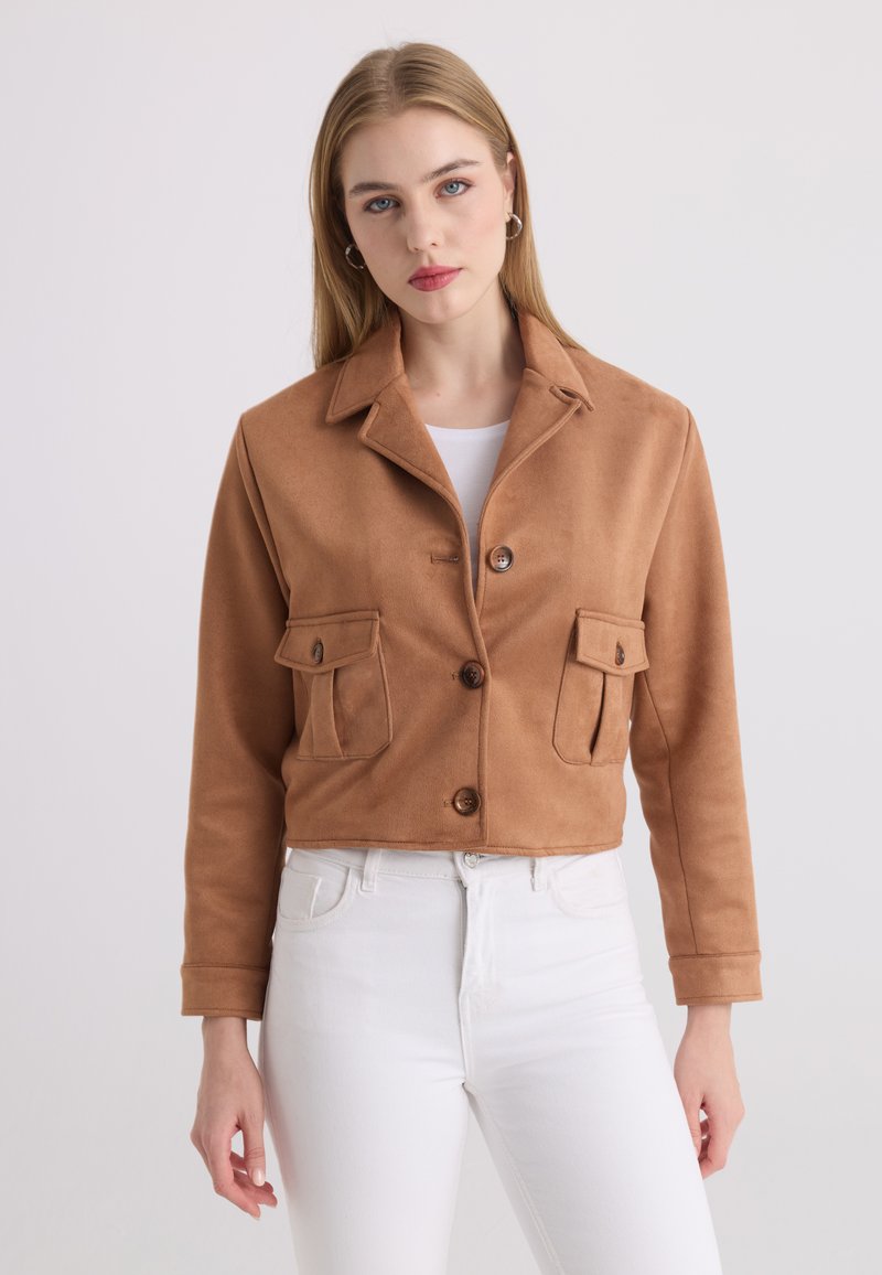Jeune femme portant une veste beige boutonnée avec poches à rabat et un pantalon blanc, debout devant un fond clair uni.