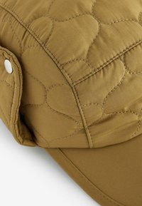 Casquette matelassée en tissu marron chaud avec des accents de motif cœur brodés. Elle présente une visière à l'avant et un détail en bouton sur le côté.