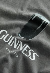 Dunkelgraues T-Shirt mit einem Motiv eines Glases Guinness und weißem Text "GUINNESS EST. 1759." Glattes Material mit subtiler Textur.