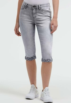 Hellgraue, gekürzte Jeans mit Leopardenmuster-Bündchen, getragen mit weißen Sneakers, gezeigt von der Taille bis zu den Waden einer stehenden Person.