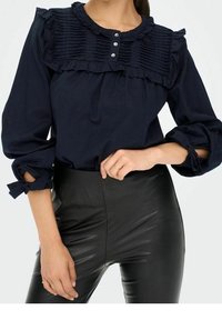 Blouse bleu marine avec un col à volants et des plis verticaux, trois boutons à l'avant, associée à un pantalon en cuir noir taille haute.