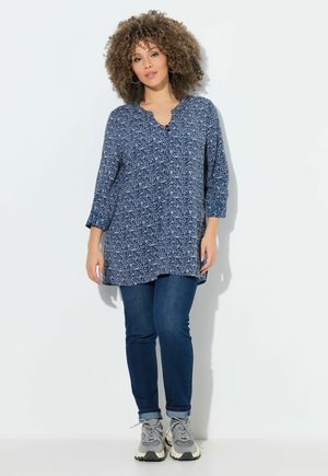 Vrouw met krullend haar, gekleed in een blauw tuniek met patroon, donkere skinny jeans en grijze sportschoenen, staand tegen een witte achtergrond.