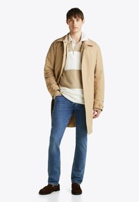 Abrigo impermeable beige sobre un suéter a rayas, jeans azules y mocasines marrones. El abrigo cuenta con botones y un corte relajado.