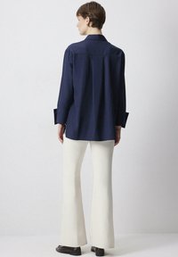 Navy blue long-sleeve shirt with a collar, featuring a relaxed fit and side slits, paired with white flared pants and dark shoes.