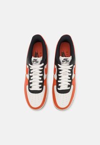 Zapatillas de cuero blancas con acentos en naranja y negro, que cuentan con una puntera perforada, cordones blancos y el clásico logo de swoosh de Nike en los laterales.
