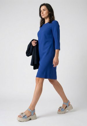 Kvinde gående i en blå kjole til knæet, holder en sort jakke, og bærer chunky lyseblå blomstrede sandaler mod en ensfarvet baggrund.