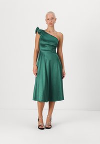 Robe en satin vert émeraude avec une épaule froncée, silhouette en A, longueur genoux et texture lisse. Des talons noires à lanières complètent le look.
