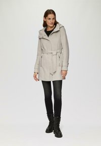 Manteau gris clair avec ceinture et capuche, doté de deux poches zippées et d'une coupe ajustée. Associé à un jean slim noir et des bottes à lacets noires.