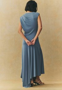 Robe longue et fluide en tissu bleu doux avec un dos drapé, présentant un ourlet asymétrique. Portée avec des talons à lanières, les mains croisées derrière.
