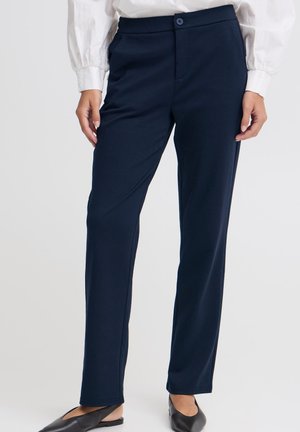Femme portant un pantalon droit bleu marine avec une fermeture à bouton et zip, associé à des chaussures plates noires et un chemisier blanc.