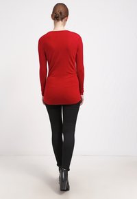 Kvinna med hår i en knut, iklädd en röd långärmad topp, svarta byxor och svarta klackade ankelboots, som går bort från en ljus enfärgad bakgrund.