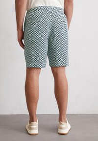 Shorts with a green and white geometric star pattern, elastic waist, back pockets with gold buttons, paired with light-colored sneakers.