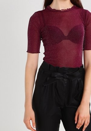 Kobieta ubrana w przezroczystą, błyszczącą bordową bluzkę z krótkimi rękawami oraz czarne spodnie z wysokim stanem, związane kokardą w talii.
