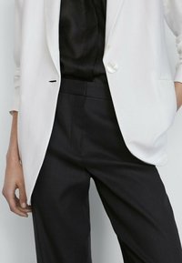 Veste blanche avec un bouton argenté unique, col à revers en encoche, et pantalon noir avec une coupe ajustée, une texture lisse, et des poches latérales.