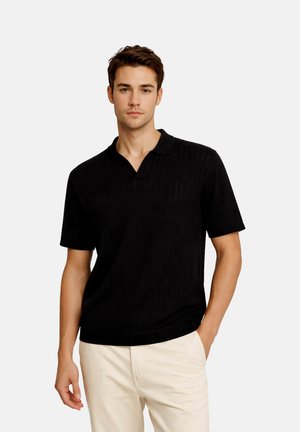 Young man wearing a black short-sleeve knit polo shirt and light beige pants, posing with one hand in pocket against a plain background.