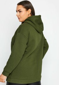 Olivgrön huvtröja med lös passform, utrustad med ribbade muddar och nederkant. Mjuk textur med en subtil logotypdetalj på ärmen.