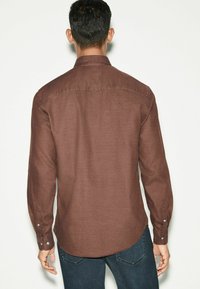 Braunes Langarmhemd mit glatter Textur, Button-Down-Kragen und abgerundetem Saum. Enthält geknöpfte Manschetten und eine Rückenpasse mit Details.