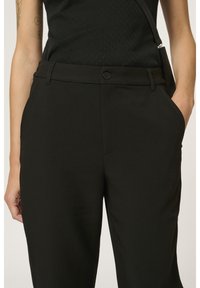 Personne portant un pantalon noir taille haute avec une fermeture à boutons et des poches latérales, associé à un haut noir sans manches et un sac bandoulière.