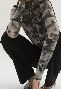 Persona accovacciata che indossa pantaloni neri, scarpe argentate e un top a maniche lunghe con motivo grigio-nero e dettagli di cucitura visibili.