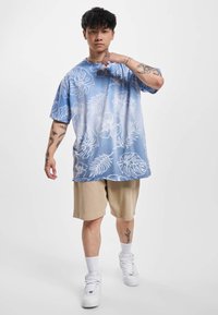Light blue oversized shirt with white floral pattern, paired with beige shorts. Model wears white sneakers and has visible tattoos on arms.