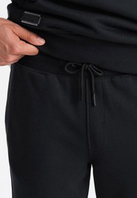 Pantalones de chándal negros con cintura ajustable, con una textura acanalada y una pequeña etiqueta de logo en la cintura. Tela suave, diseño ajustado.