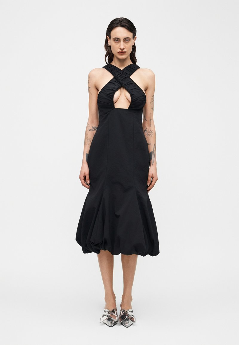 Femme portant une robe midi noire sans manches avec un col halter croisé froncé et un ourlet bouffant, associée à des chaussures pointues argentées.