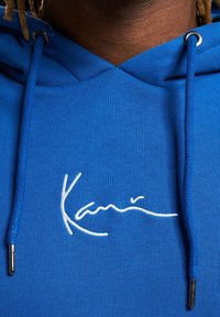Sudadera con capucha azul hecha de algodón con una textura suave. Presenta un logotipo blanco bordado en cursiva y puntas metálicas en los cordones.